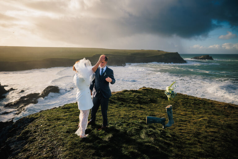 Image showing wedding couple during their winter wedding standing on the headland with the sea behind them. the brides veil is billowing in the wind and she has thrown her wellies and flowers to her side. Captured by Cornwall Wedding Photographer Chris Armstrong Photography