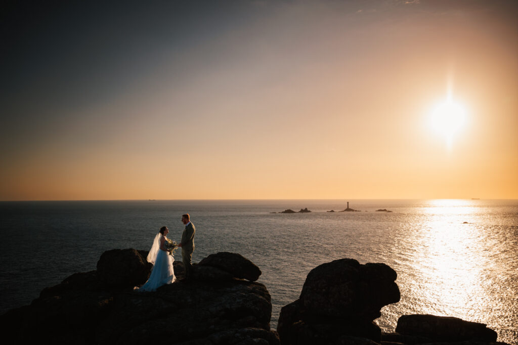 Just married couple standing on the rocks at sunset after the cliff-top wedding at Lands end in Cornwall captured by cornwall wedding photographer Chris Armstrong Photography